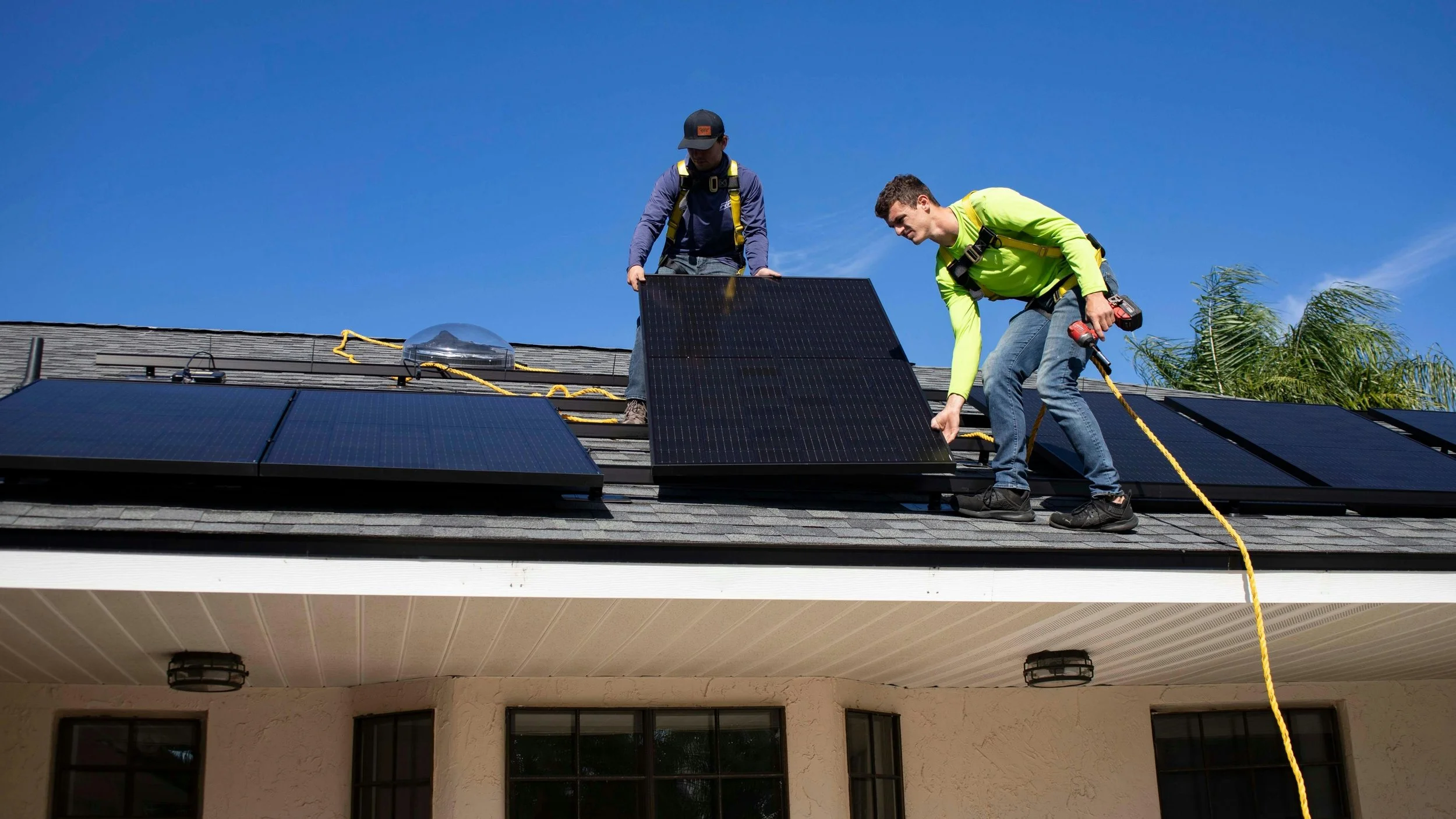 Two workers installing solar panels on a roof under a clear blue sky.