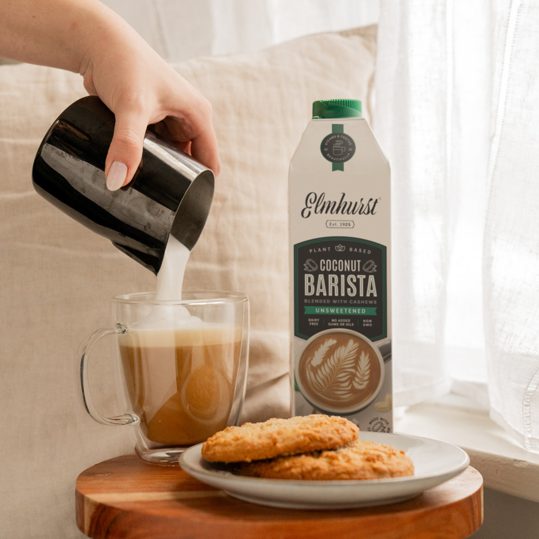 Hand pouring Elmhurst Coconut Barista plant-based milk into a glass mug of coffee beside a carton of the product, with two cookies on a plate on a small wooden table by a bright window.