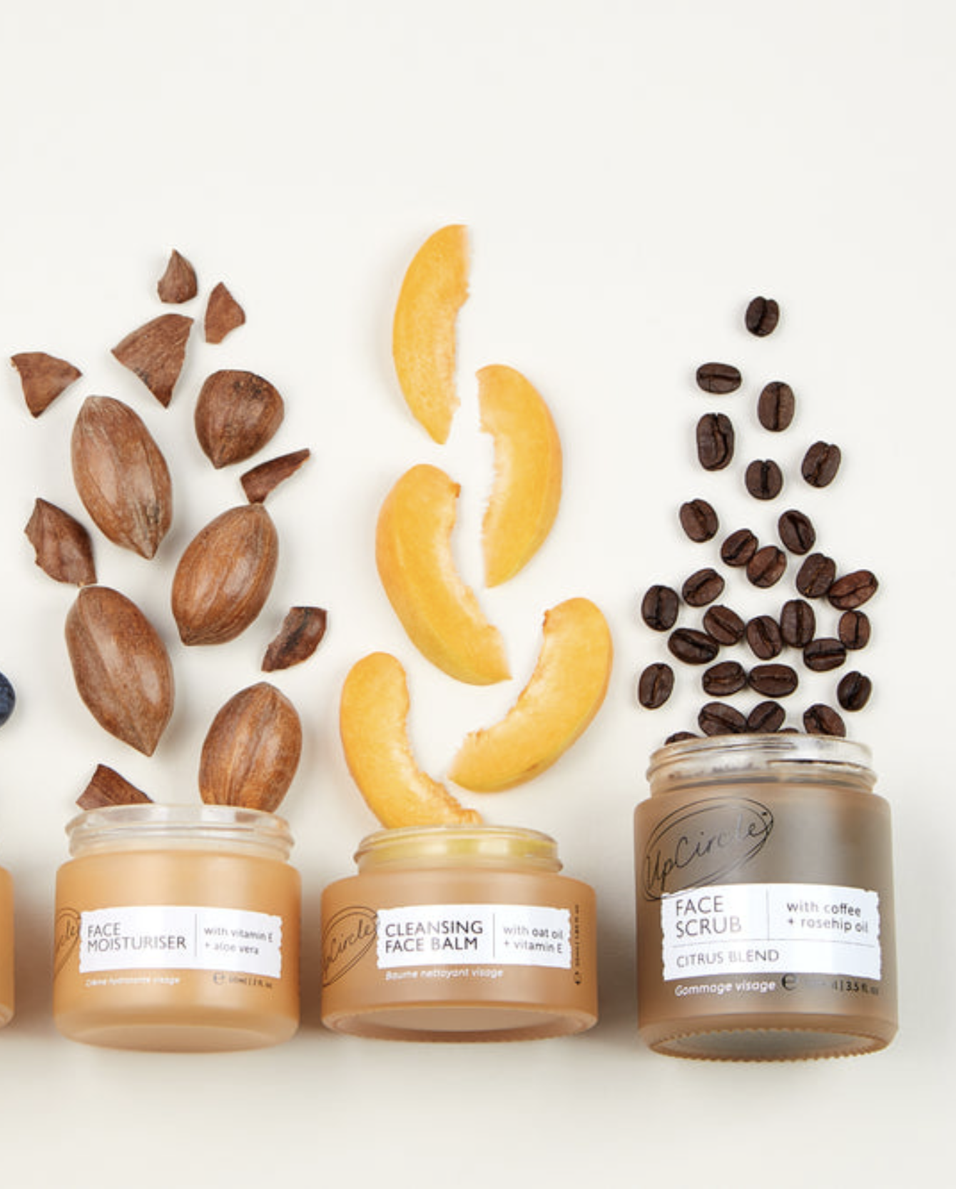 Flat lay of UpCircle skincare jars including face moisturizer, cleansing face balm and citrus blend face scrub arranged beneath almonds, orange slices and coffee beans representing upcycled natural ingredients