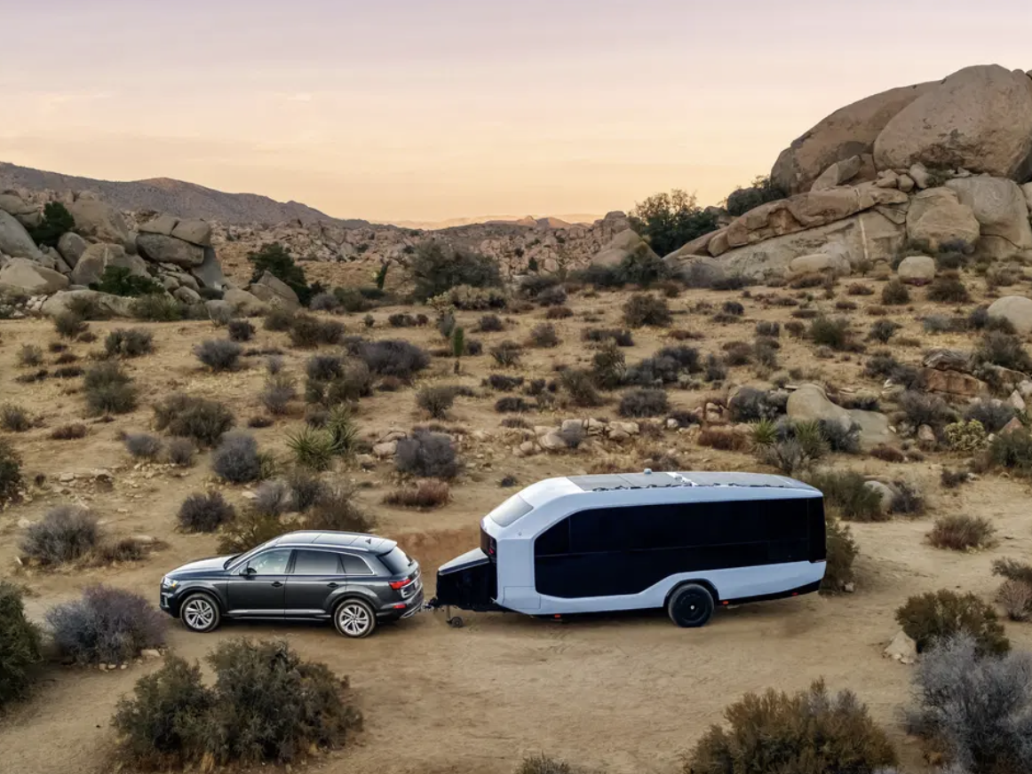 The Pebble Flow electric RV trailer being towed through a desert landscape at sunset, with rocky outcroppings and desert scrub in the background.