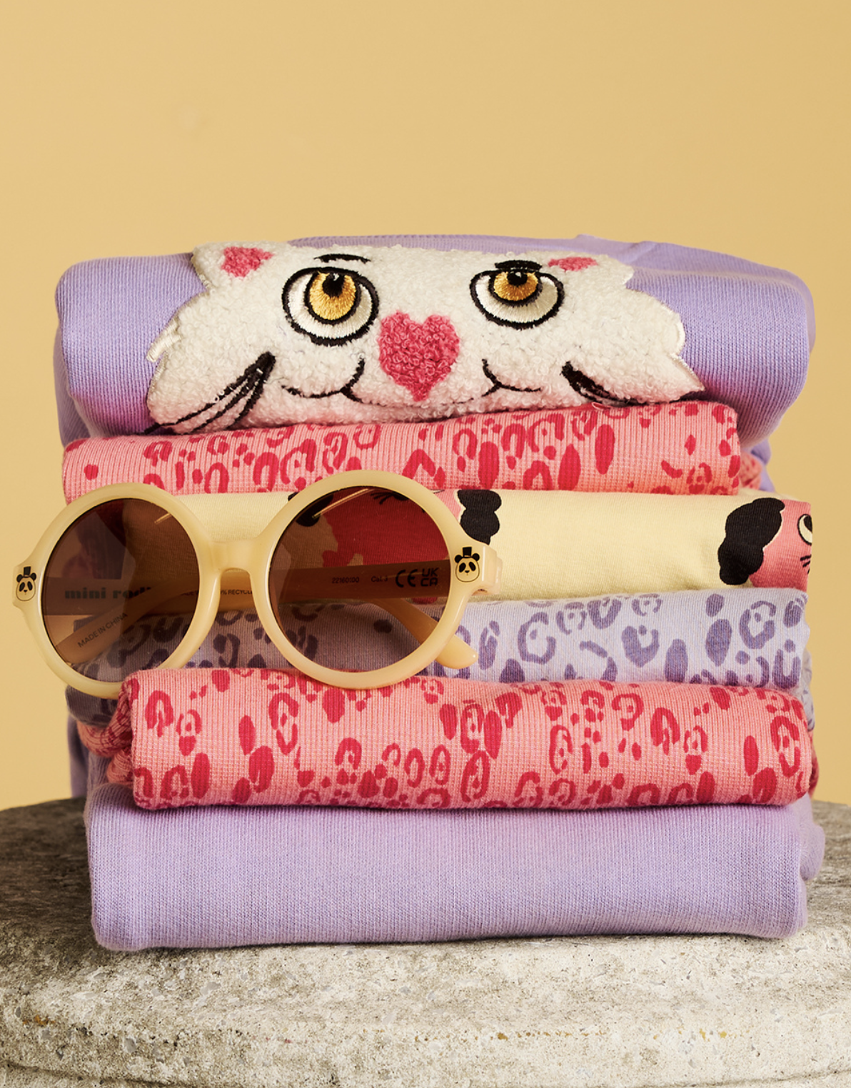 Folded stack of colorful Mini Rodini clothing featuring playful animal graphics, pink and lavender prints and round cream-colored sunglasses on top