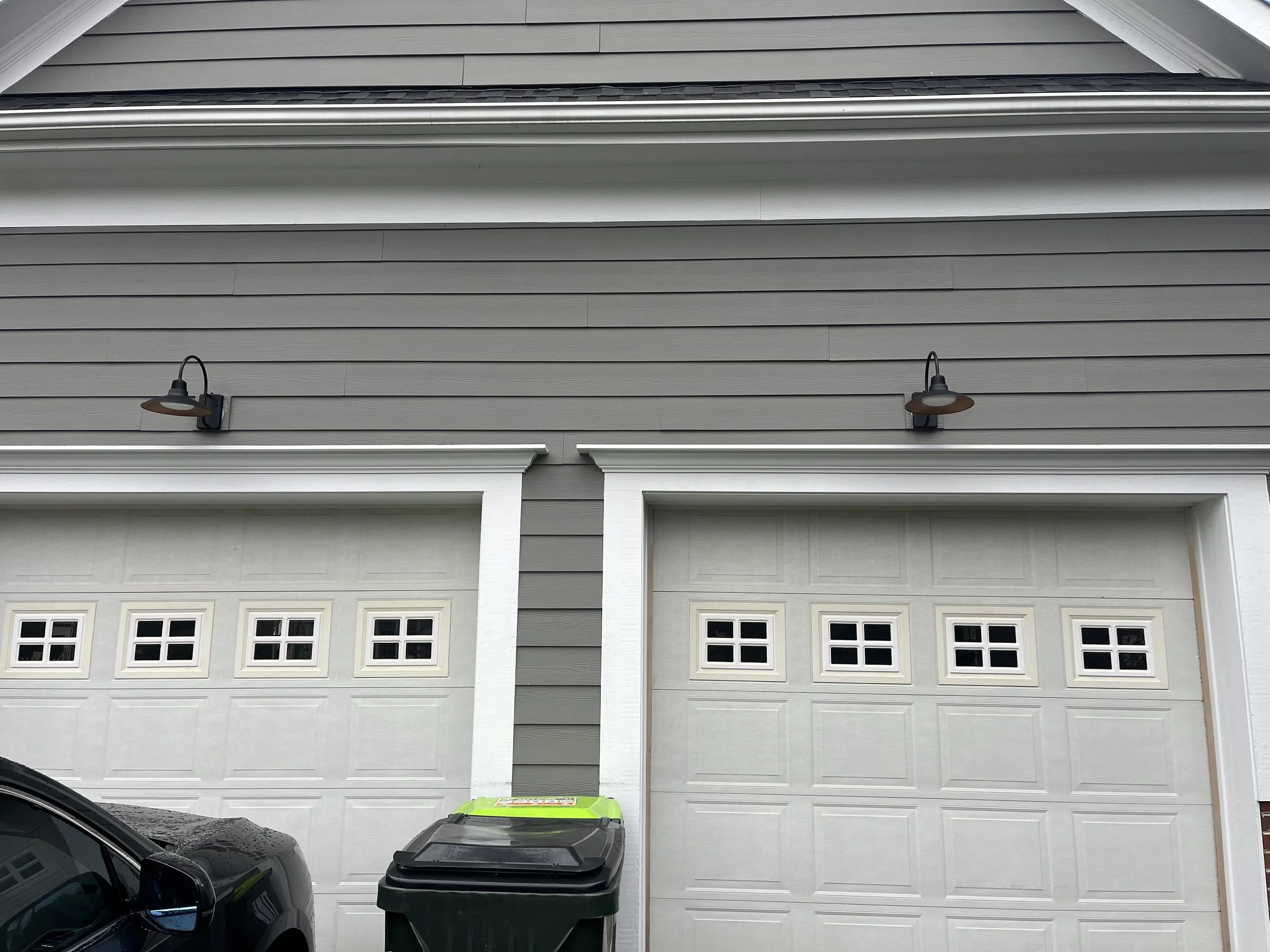 Two garage doors with small square windows at the top, grey siding, black outdoor lights, a green and black trash bin, and a portion of a parked car in front.