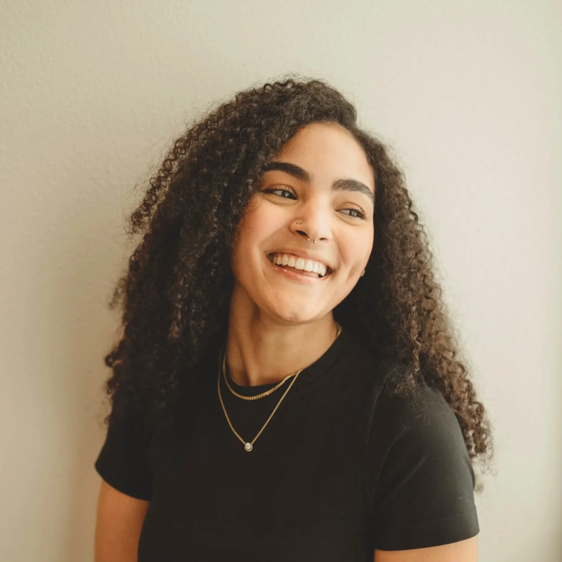 A young woman with curly dark hair smiling, wearing a white knitted sweater and jewelry, standing against a plain white wall.