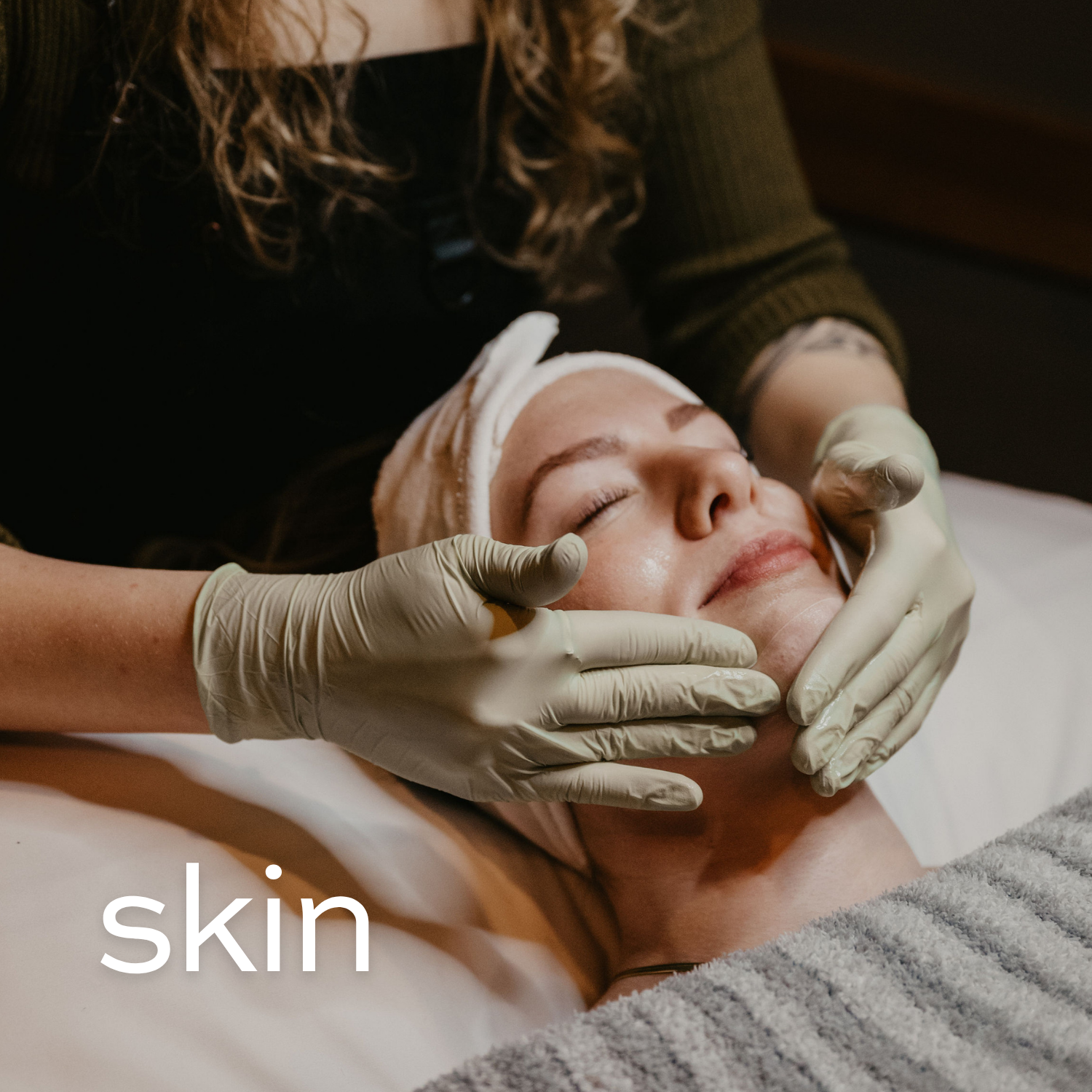 Aesthetician performing a facial treatment on a client lying down with a headband, in a spa setting.