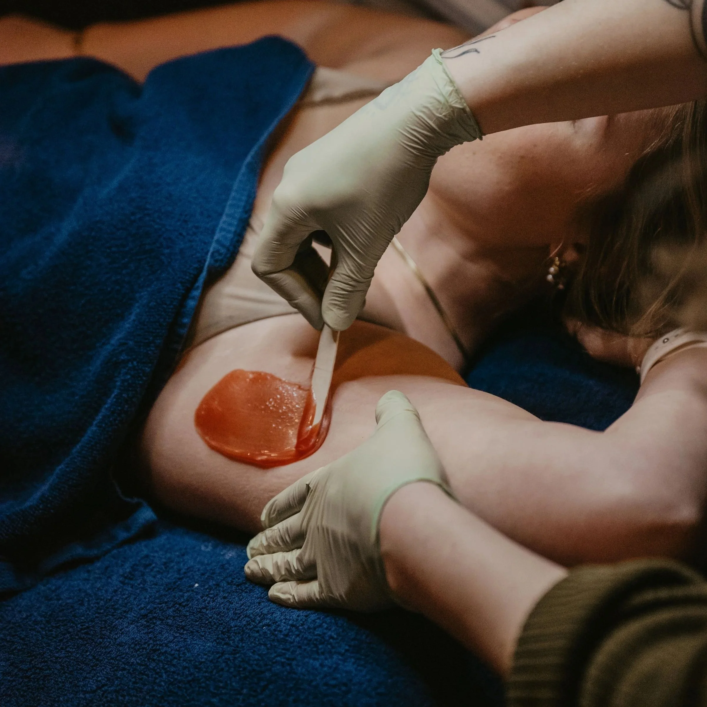 Person receiving a waxing treatment on their body with a spatula and wax applied, wearing gloves, and partially covered by a blue towel.