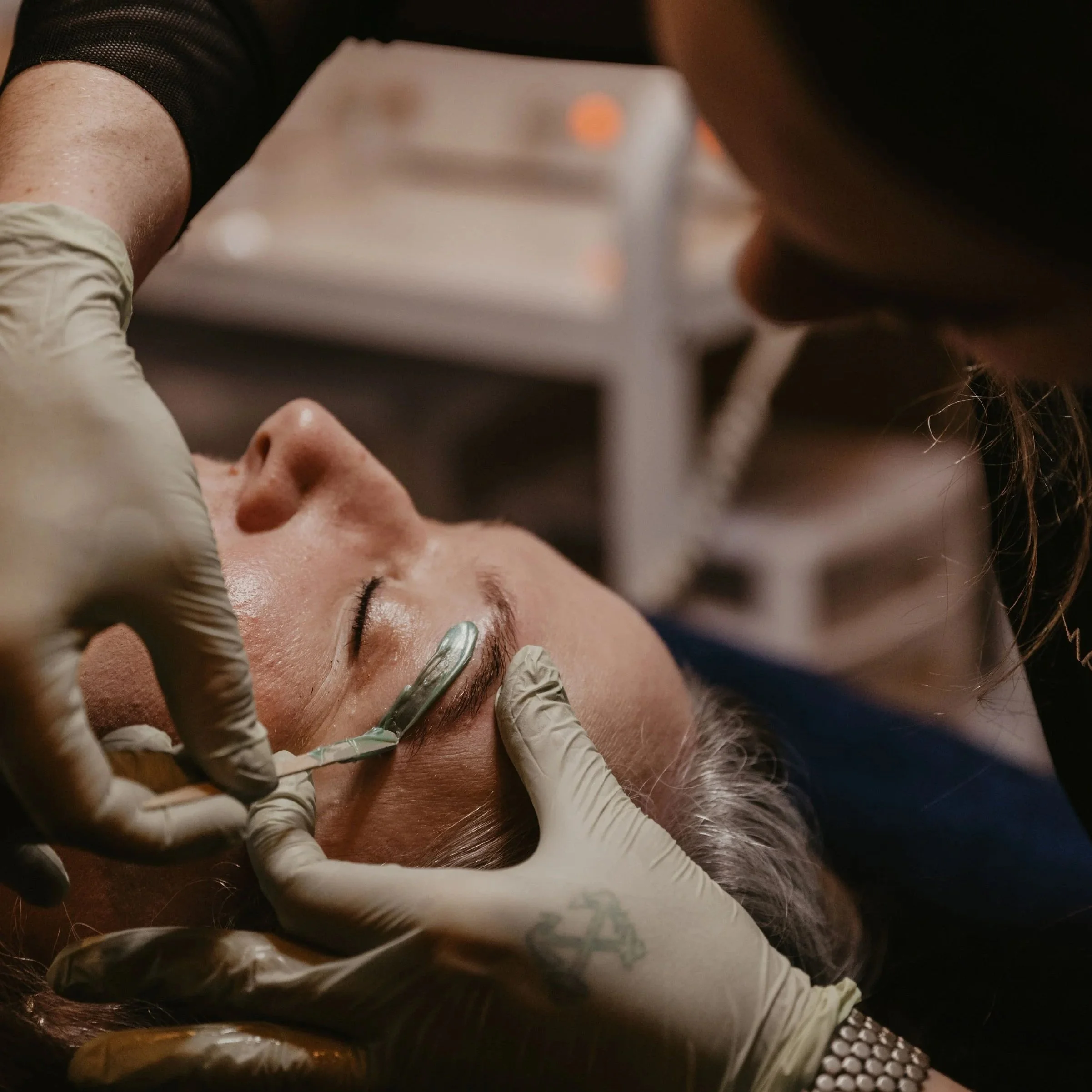 A person applying beauty treatment to another person's eyebrows, using gloved hands and a tool. The recipient is lying down with closed eyes.