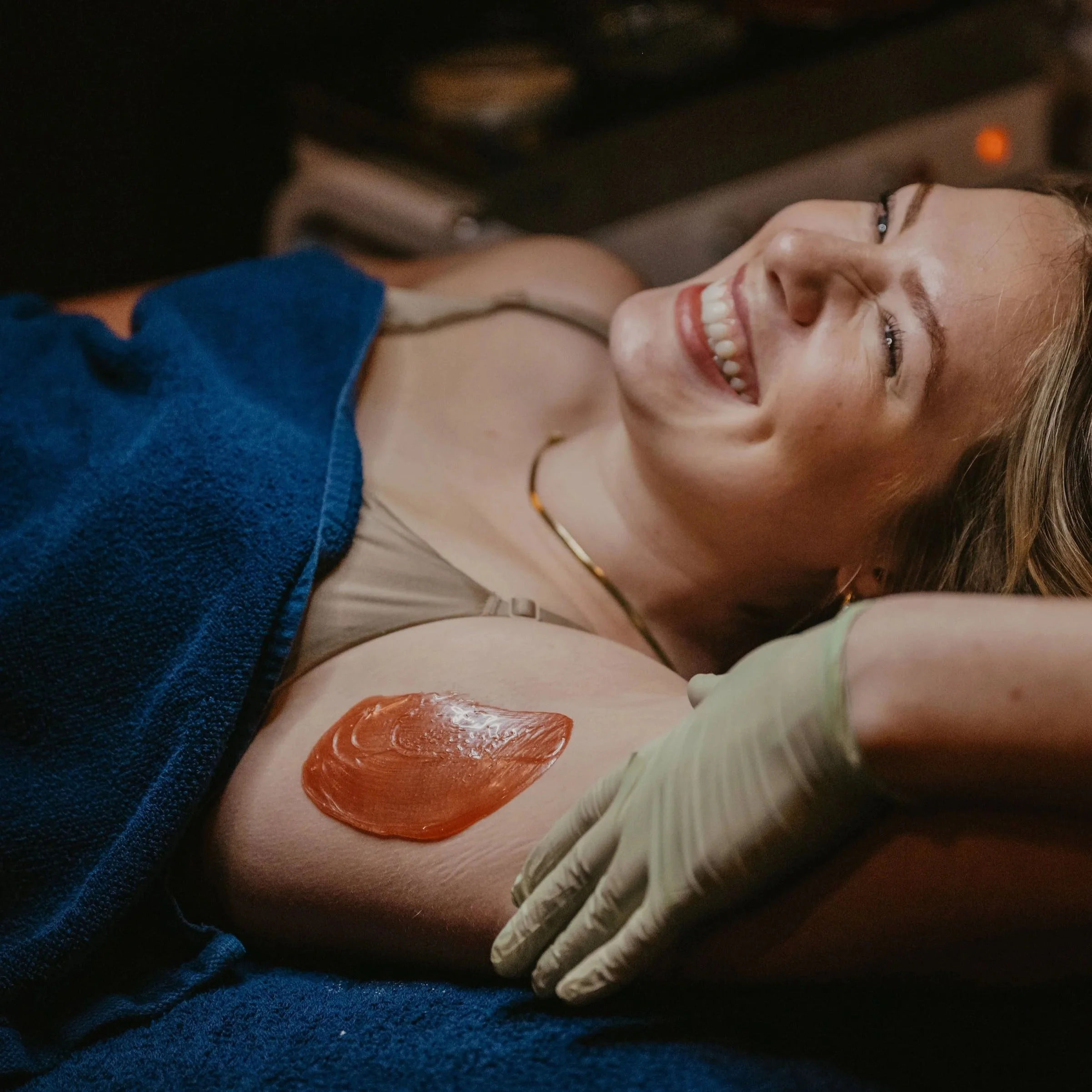 Person receiving an armpit waxing treatment, smiling, with wax applied on the skin; lying on a blue towel; gloved hand nearby.