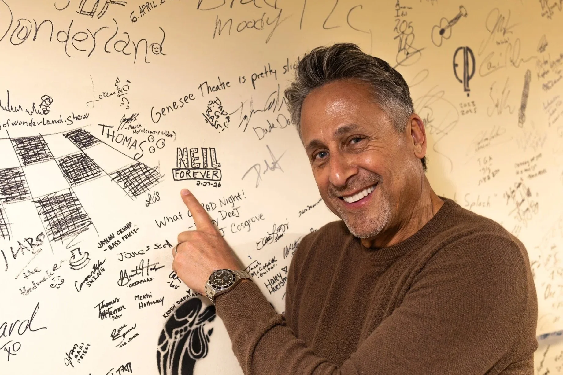 A smiling middle-aged man with gray hair points at a wall covered with signatures, drawings, and handwritten messages.