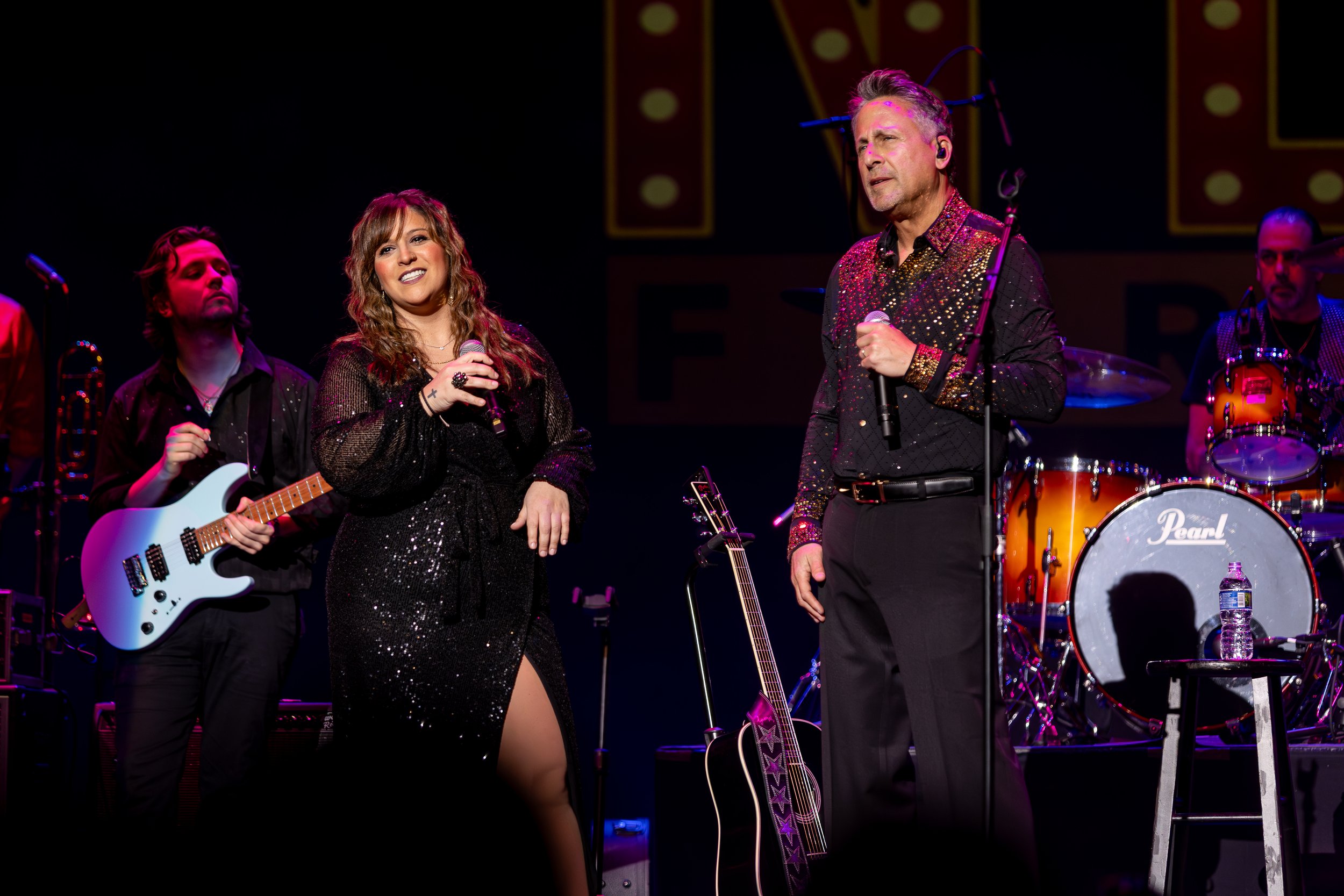 A singer and a male performer with a microphone on stage, accompanied by band members playing guitar and drums in a concert setting. The stage has colorful lighting and musical equipment.