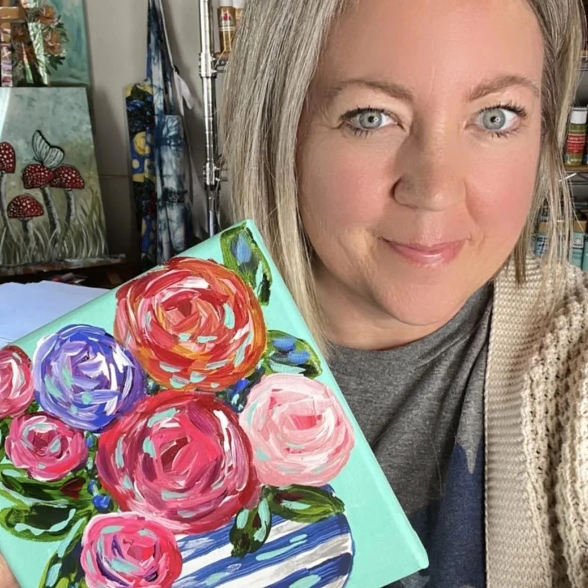 A woman with blonde hair and blue eyes holding a colorful floral painting with roses in a vase.