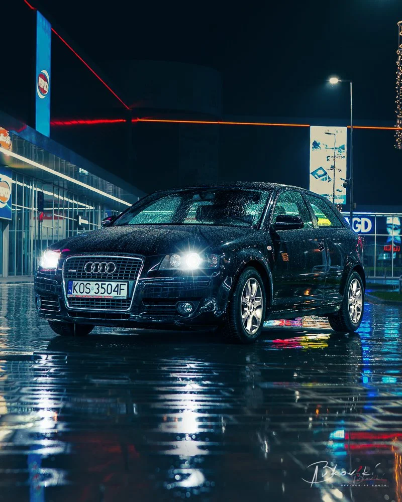 Black Audi A3 parked on wet pavement at night, illuminated by streetlights and nearby signs, with reflections on the ground.