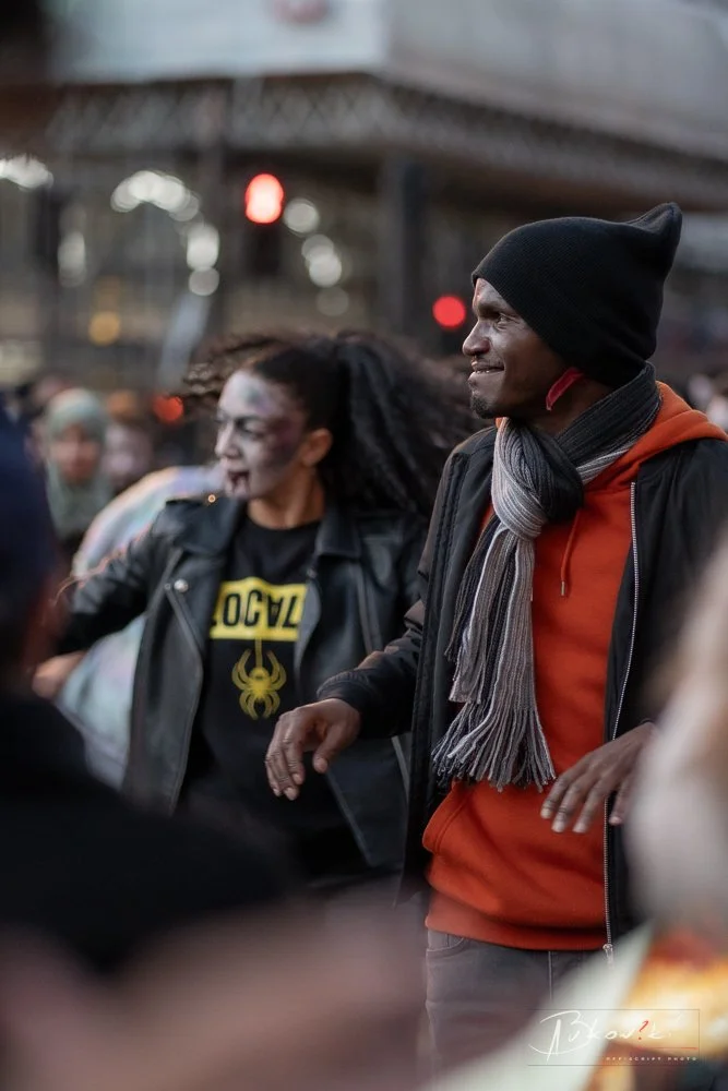 A man and woman smiling and enjoying themselves outdoors in a crowd near a bridge, with the woman dressed in a leather jacket and the man wearing a beanie, scarf, orange hoodie, and black jacket.