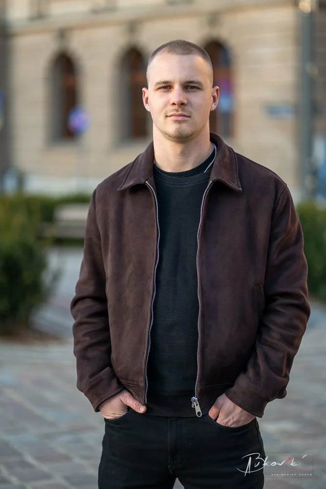 A young man standing outdoors in front of a blurred building, wearing a brown zip-up jacket over a black shirt, with hands in his pockets, looking at the camera.