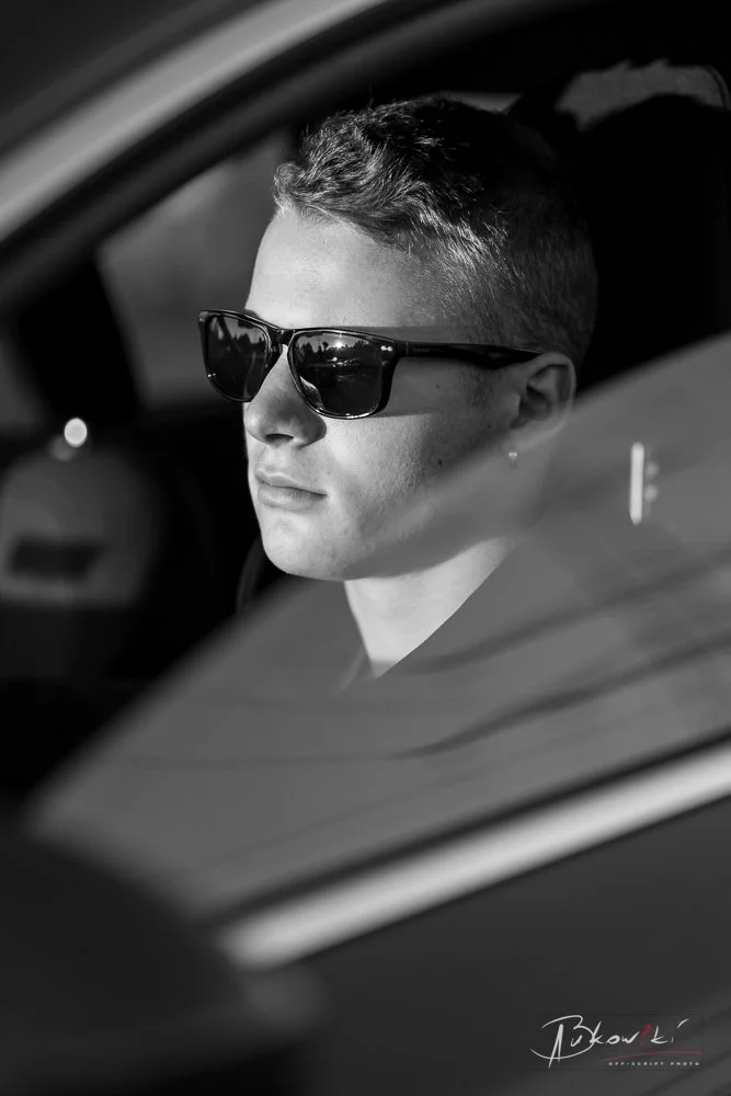 A person with short hair and sunglasses sitting in a car, seen through the window, in black and white.