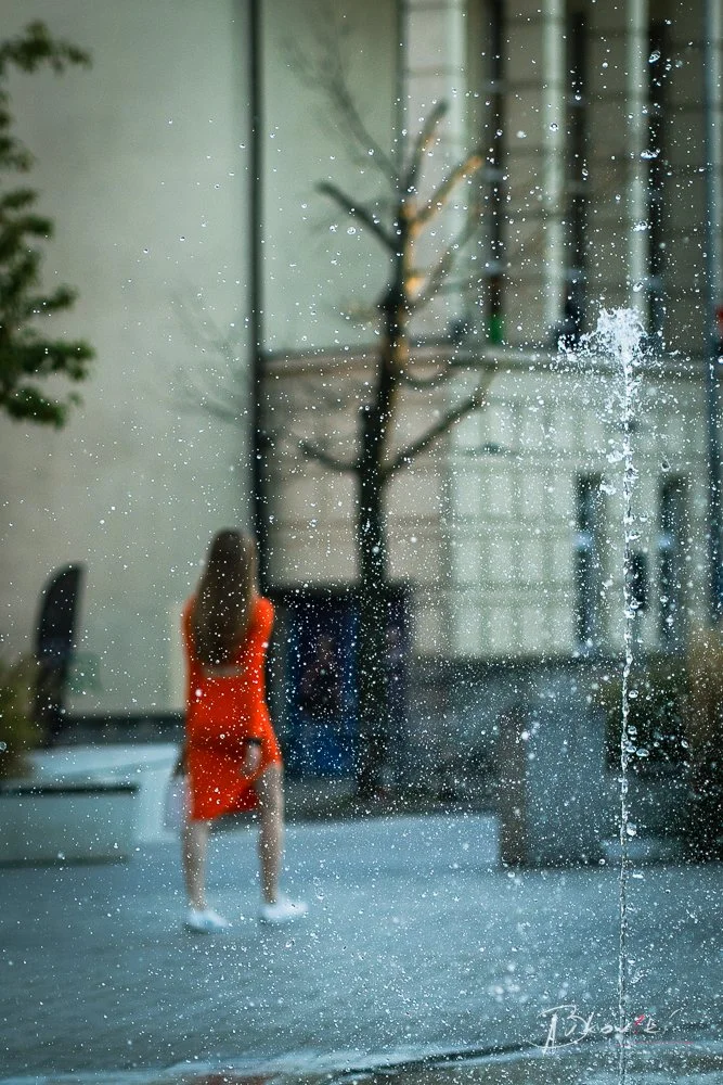 A woman in a red dress standing outdoors behind a glass window with water droplets, a leafless tree, and buildings in the background.