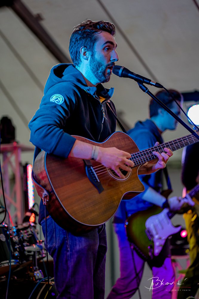 Man playing a wooden acoustic guitar and singing into a microphone during a live performance under a tent at night.