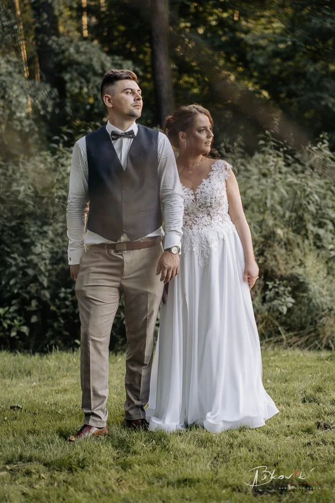 A groom and bride standing outdoors in a wooded area, holding hands, with trees and foliage in the background, dressed in formal wedding attire.