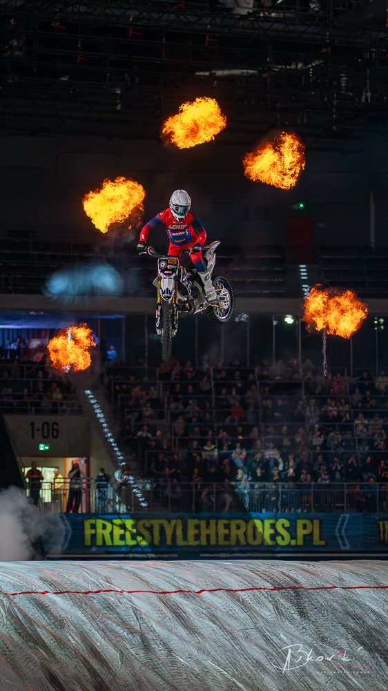 Freestyle motocross rider mid-air with fire explosions around during an indoor stunt show, audience watching from stands.