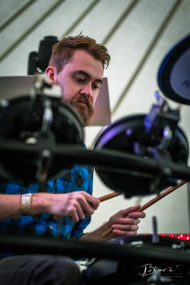 A man with a beard and plaid shirt playing drums at a music event.
