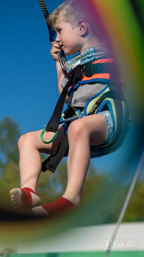 A young boy with light-colored hair and fair skin riding a zip line during daytime, wearing a harness and comfortable clothing, with a clear blue sky and some trees in the background.