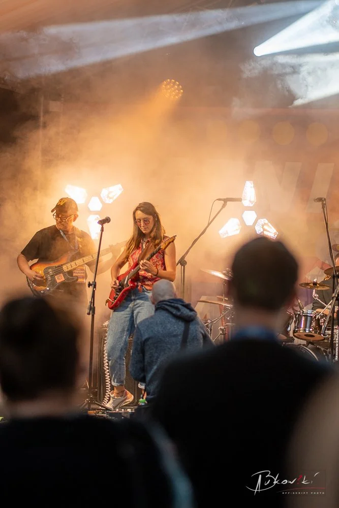 Musicians performing on stage with guitars, surrounded by stage lights and fog, audience visible in foreground.