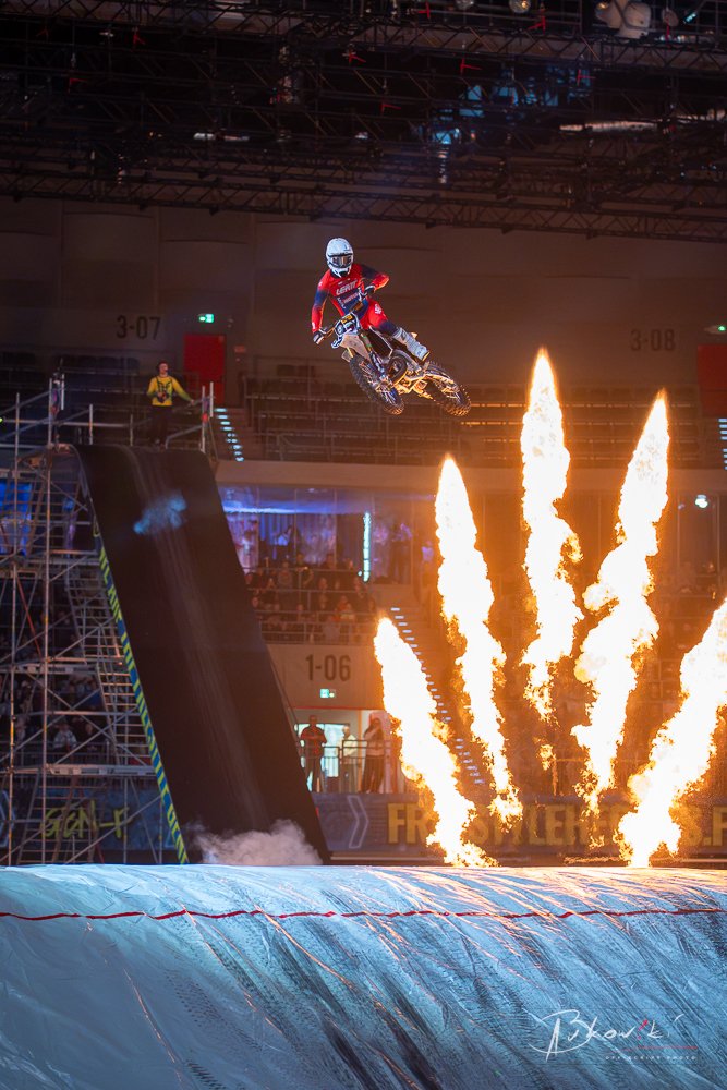A motocross rider in a red suit and white helmet performing a jump over fire during an indoor event, with a large ramp and flames on the ground.