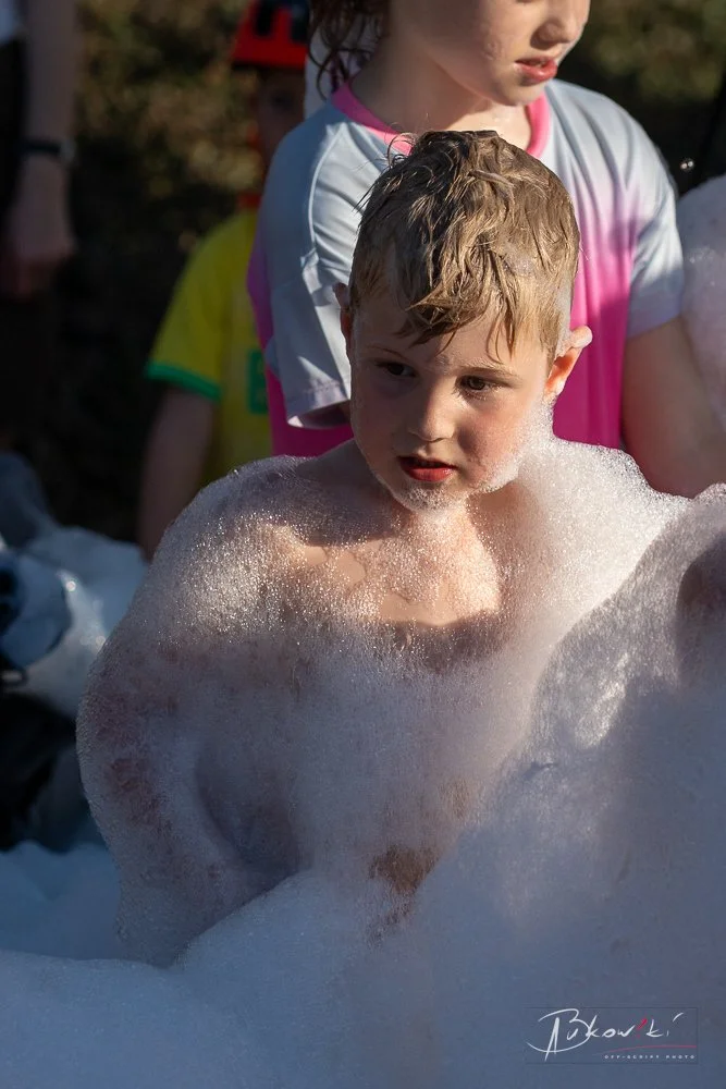 A young boy with wet hair and foam on his shoulders and chest, surrounded by bubbles, participating in a foam event outdoors. A girl with dark hair and a pink and white shirt stands behind him.