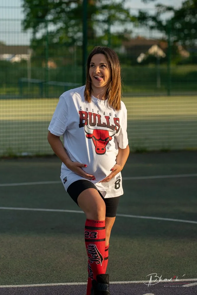 A woman in a Chicago Bulls shirt and red sports socks is standing on a tennis or basketball court, smiling and holding her pregnant belly.