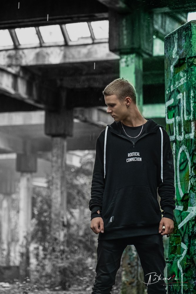 A young man with blonde hair stands under a bridge, wearing a black hoodie with 'Rootical Connection' written on it, black pants, and a necklace. The background features graffiti and urban decay.