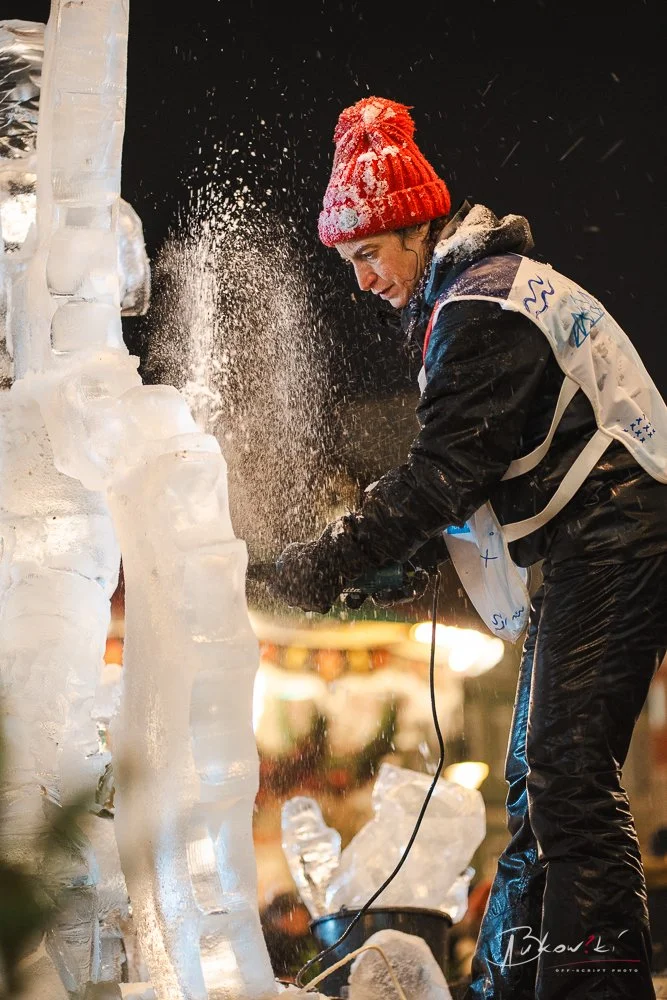 Person wearing a red knit hat and winter clothing carving ice with a power tool, creating ice chips and spray.