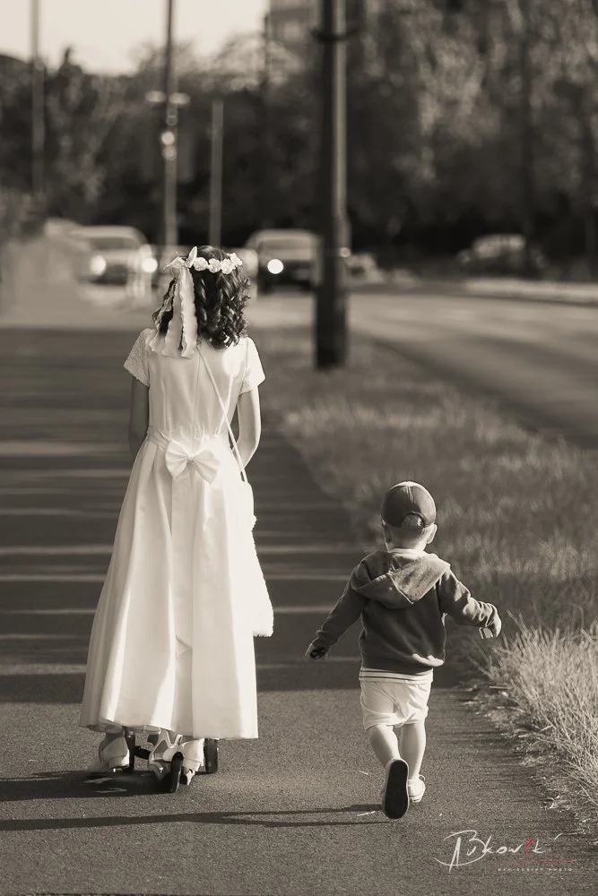 A girl in a white dress with a flower crown riding a skateboard, walking alongside a young boy in a hoodie and shorts on a sidewalk during daytime.