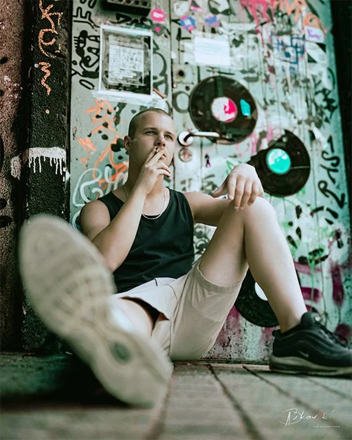Young woman sitting against a graffiti-covered wall with speakers, wearing a black tank top, beige shorts, and sneakers, appearing to be lost in thought.