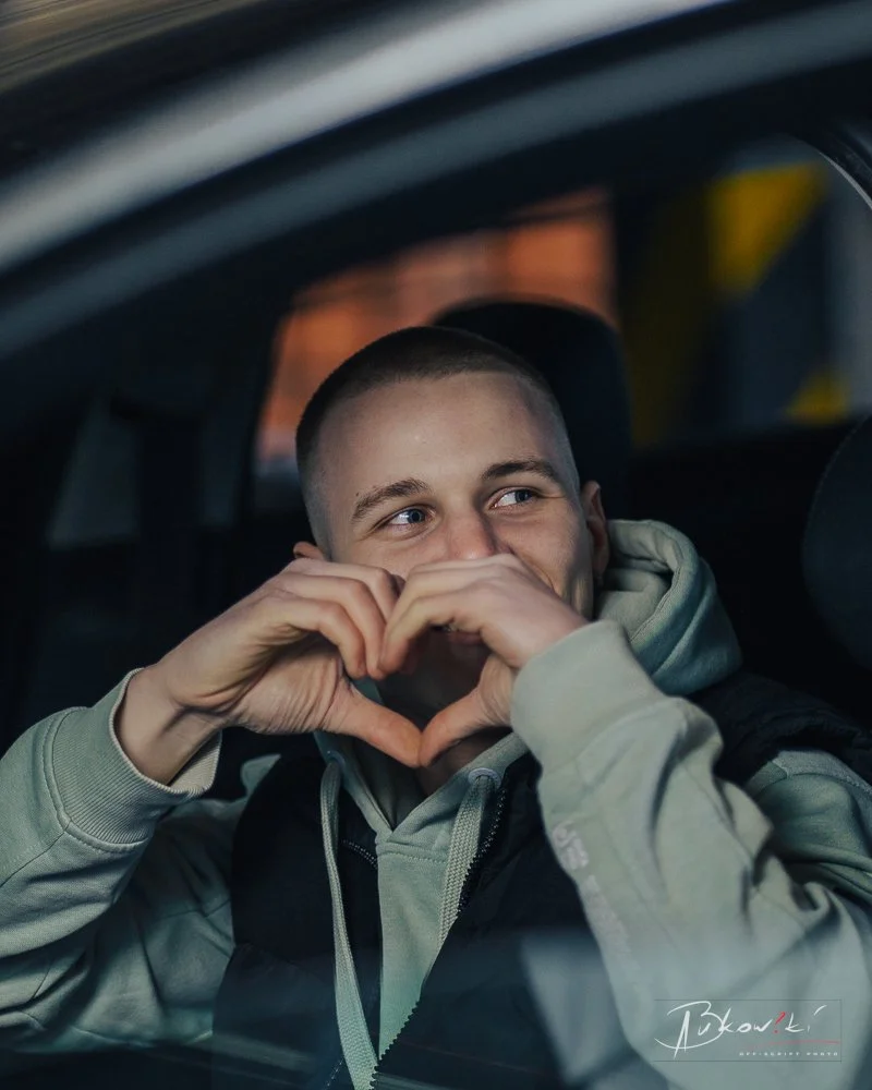 A young man with short hair sitting in a car, smiling and making a heart shape with his hands near his face.