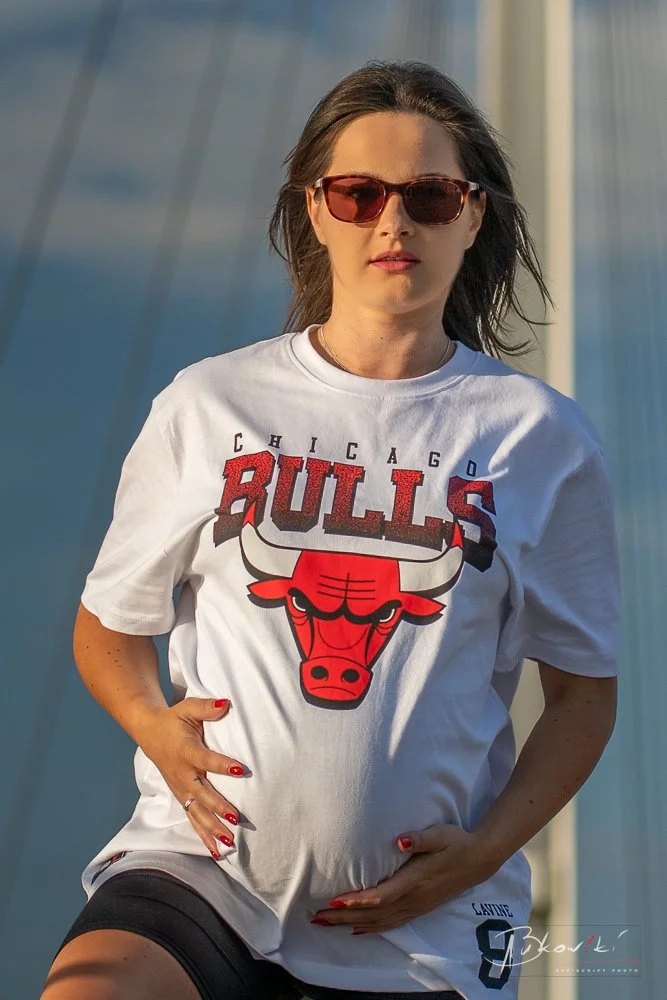 A woman with brown hair wearing sunglasses and a Chicago Bulls t-shirt, standing outdoors on a bridge with blue sky background.