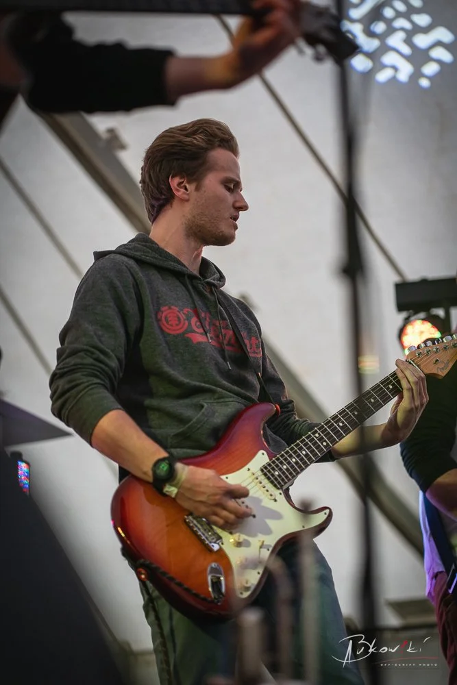Young man playing an electric guitar on stage under a tent, wearing a dark hoodie and a watch.