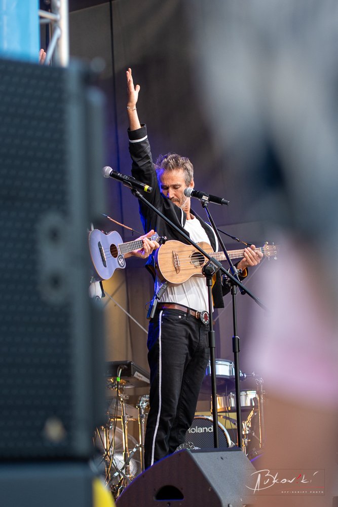 A man with gray hair and a beard playing an acoustic guitar on stage, raising his right hand, with microphones and musical equipment around him, during a live performance.