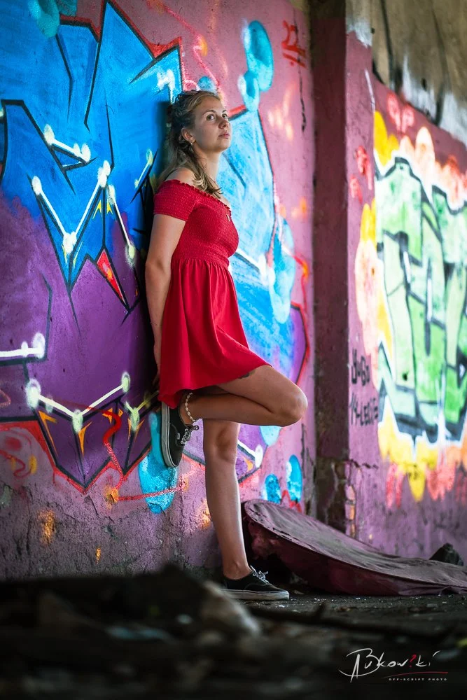 A young woman in a red dress and black shoes leans against a colorful graffiti-covered wall, standing on one leg with her other leg bent and foot resting on the wall.