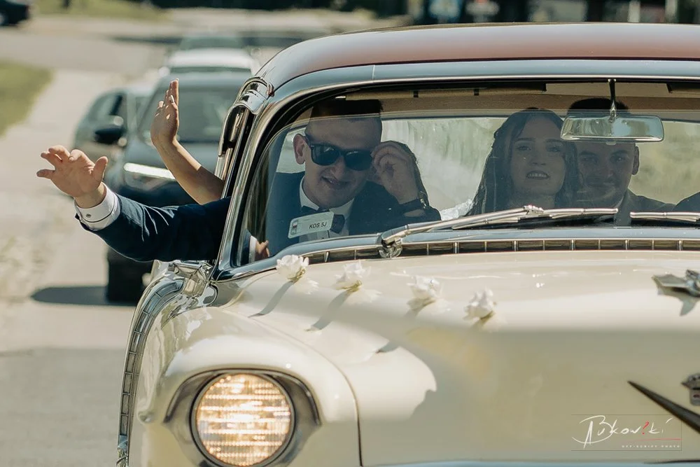 A wedding limousine decorated with white flowers carrying four people, including a bride in a white dress and veil, and three men in the front seating, one of whom is waving out the window.