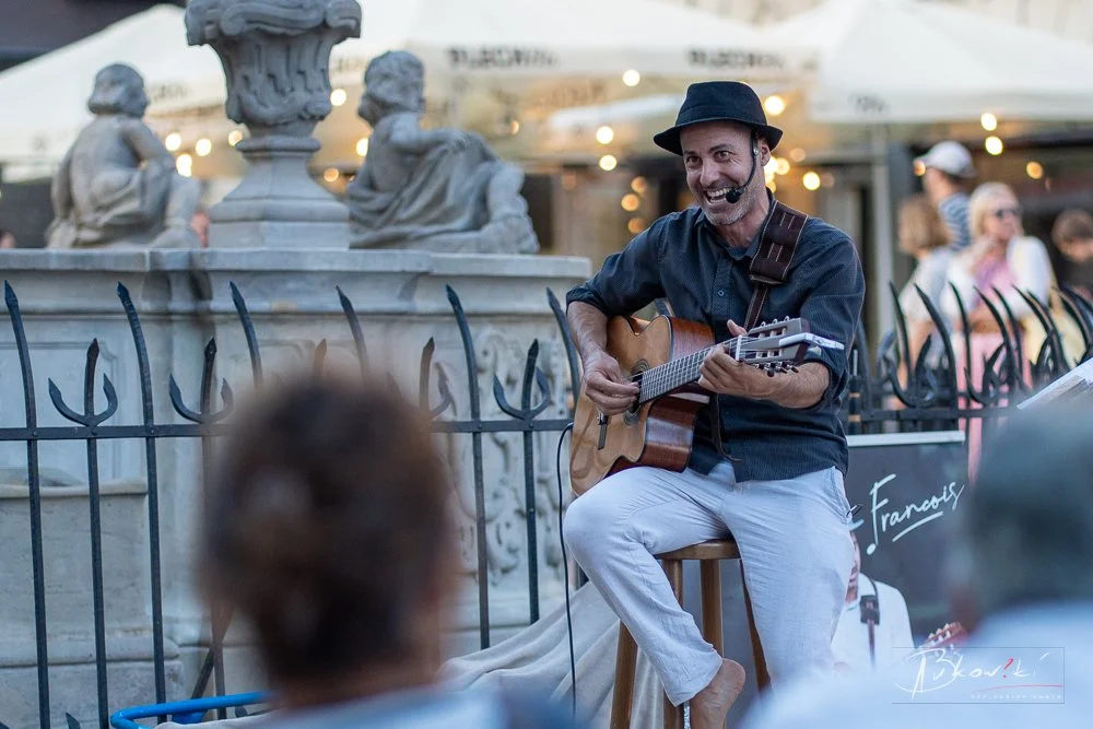 Street musician playing guitar and singing with a microphone in an outdoor setting, surrounded by people and decorative statues.