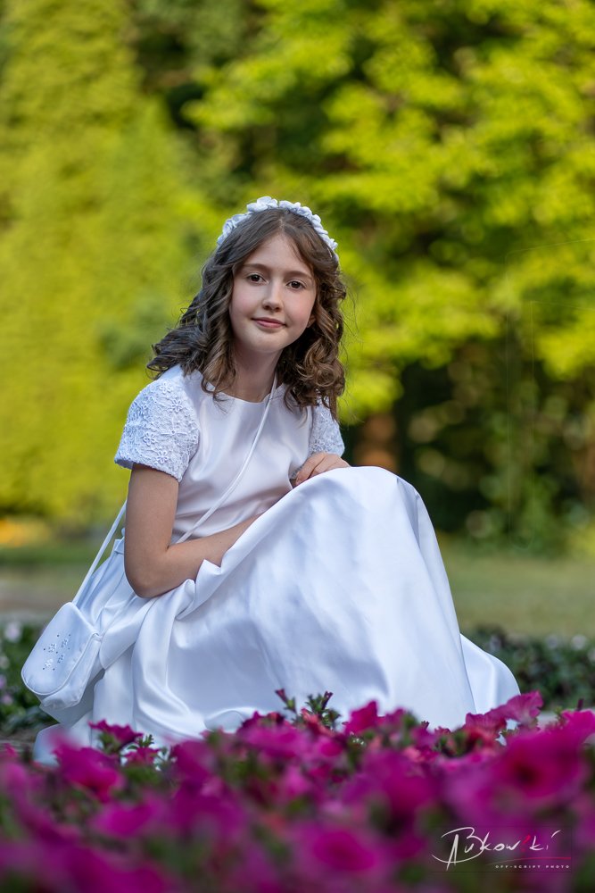 Young girl in a white dress and lace sleeves sitting outdoors among pink flowers, with a blurred green background.
