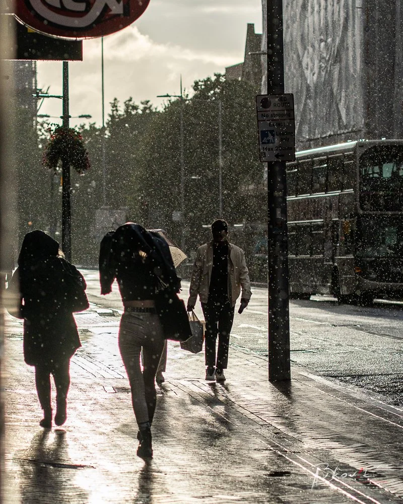 People walking on a rainy city sidewalk with sunlight and rain droplets in the air, street signs, and a bus in the background.