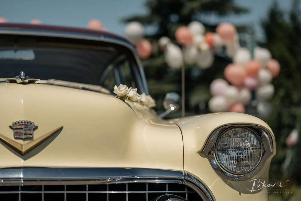 Close-up of a vintage cream-colored Cadillac car, with part of the front grille and headlight visible, and decorative flowers on the hood, with a background of blurred pastel balloons and trees.