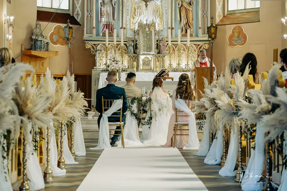 A wedding ceremony taking place in a church with a priest at the altar, couples seated facing the altar, floral arrangements along the aisle, and wedding guests seated on both sides.
