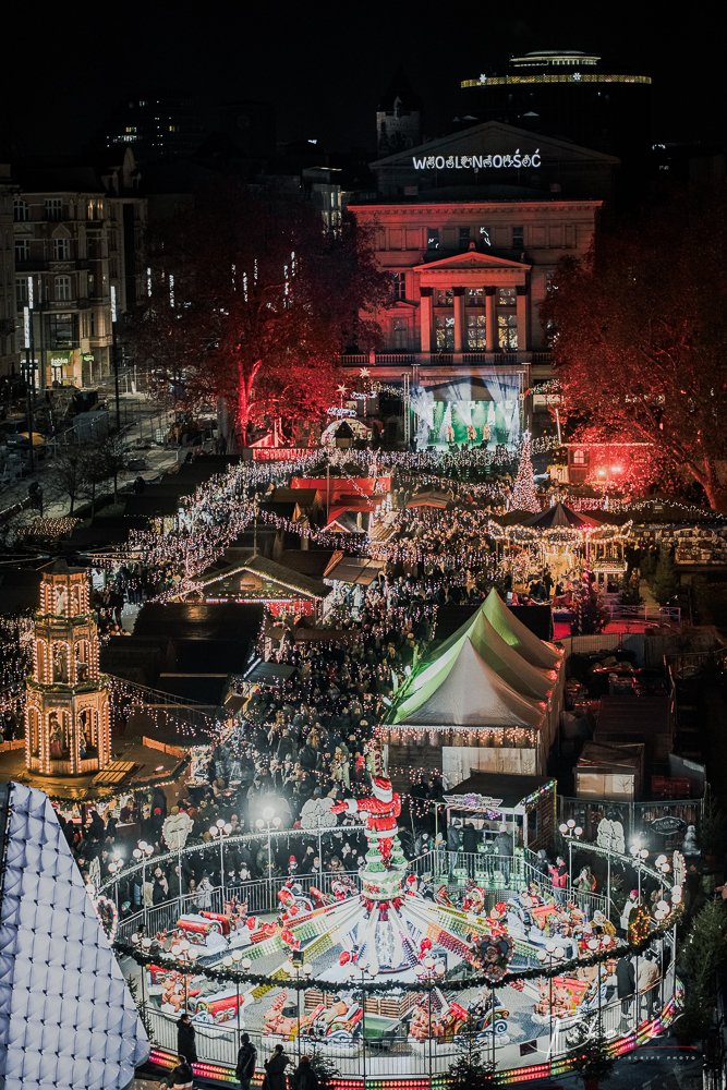 Night view of a Christmas market filled with Christmas lights, decorated tents, and a carousel with Santa Claus decorations in the foreground, with a large building and trees illuminated in red in the background.
