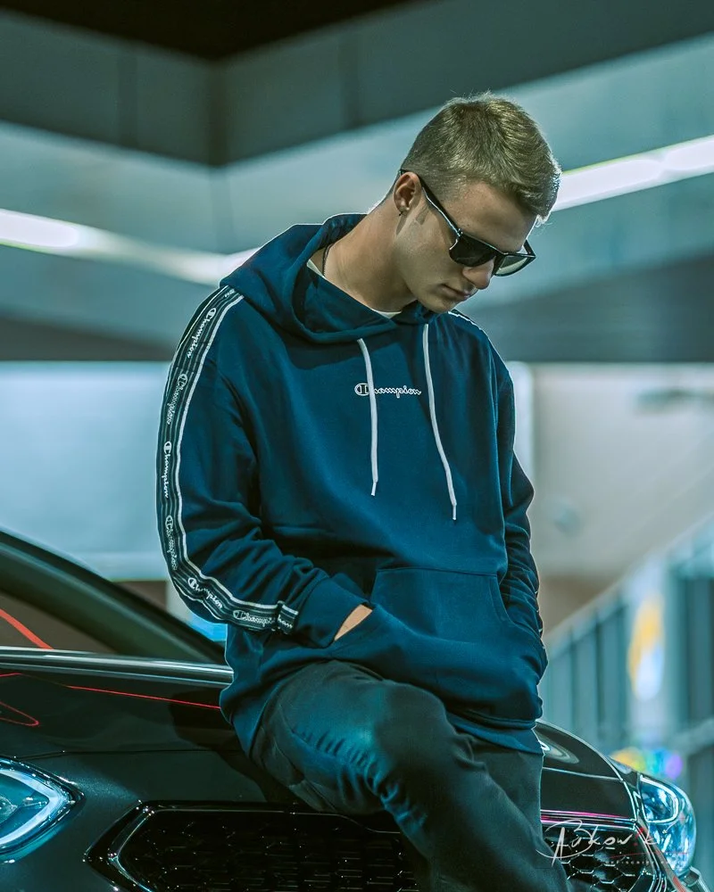 A young man wearing sunglasses, a navy blue Champion hoodie, and black pants, sitting on the hood of a black car in an indoor parking garage.
