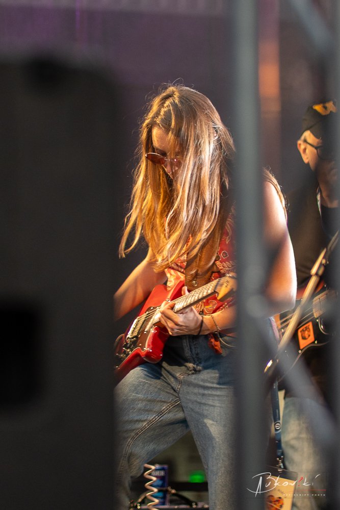 A woman playing an electric guitar during a live music performance, wearing sunglasses, a sleeveless top, and jeans, with stage equipment visible.