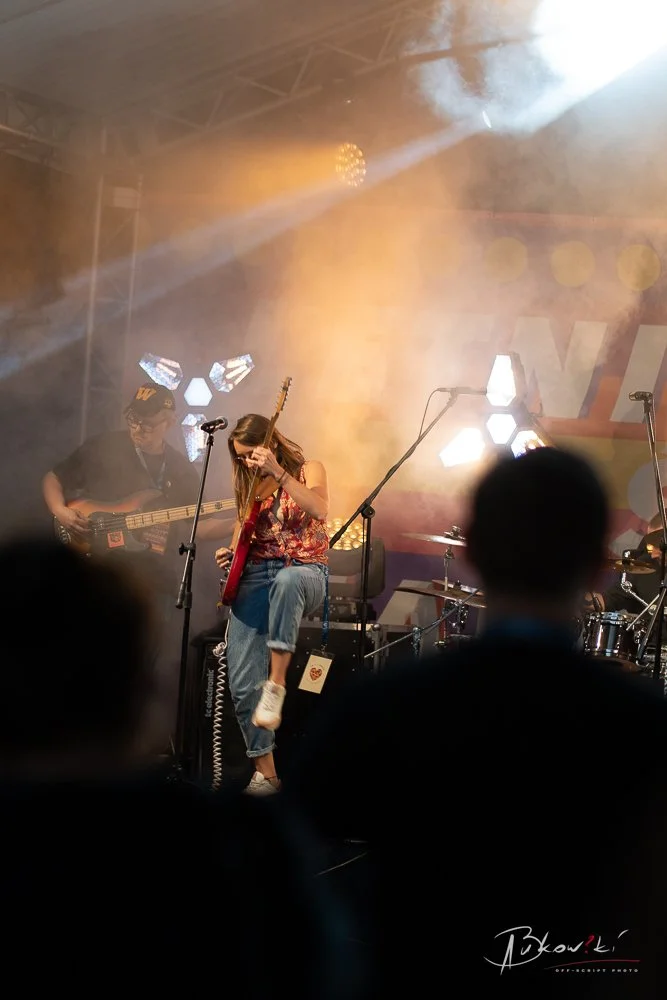 Female guitarist on stage performing with smoke and bright stage lights, accompanied by a bassist in the background, in a live concert setting with an audience in the foreground.