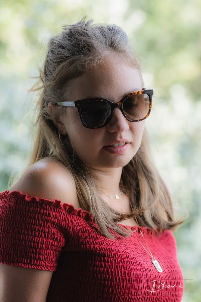 A girl wearing sunglasses and a red off-shoulder top, with a blurred outdoor background.