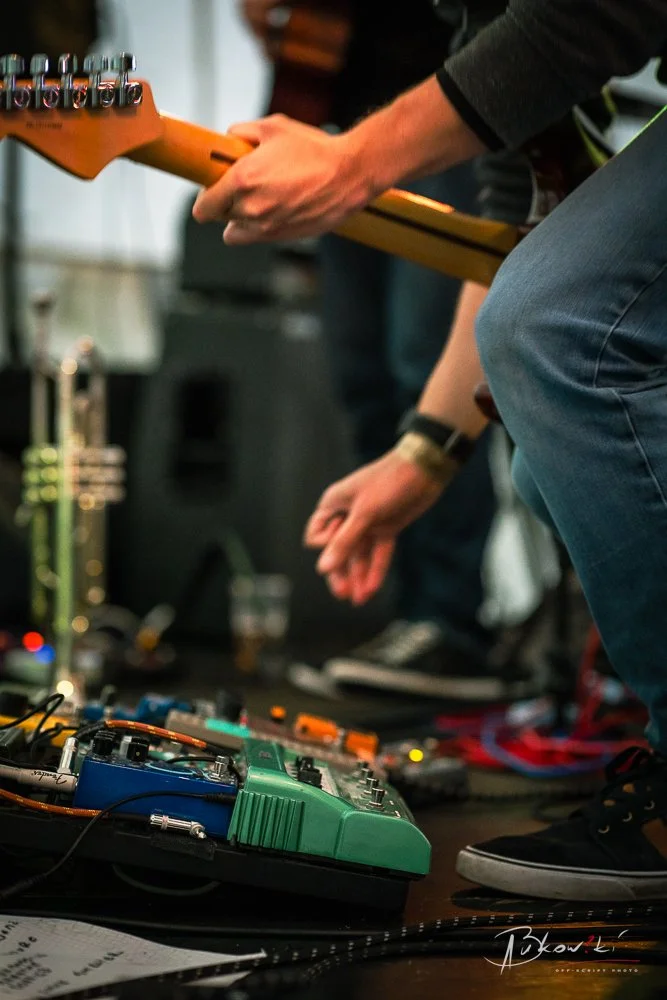 Close-up of a musician's hands and feet as they play an electric guitar and operate effects pedals on a stage.