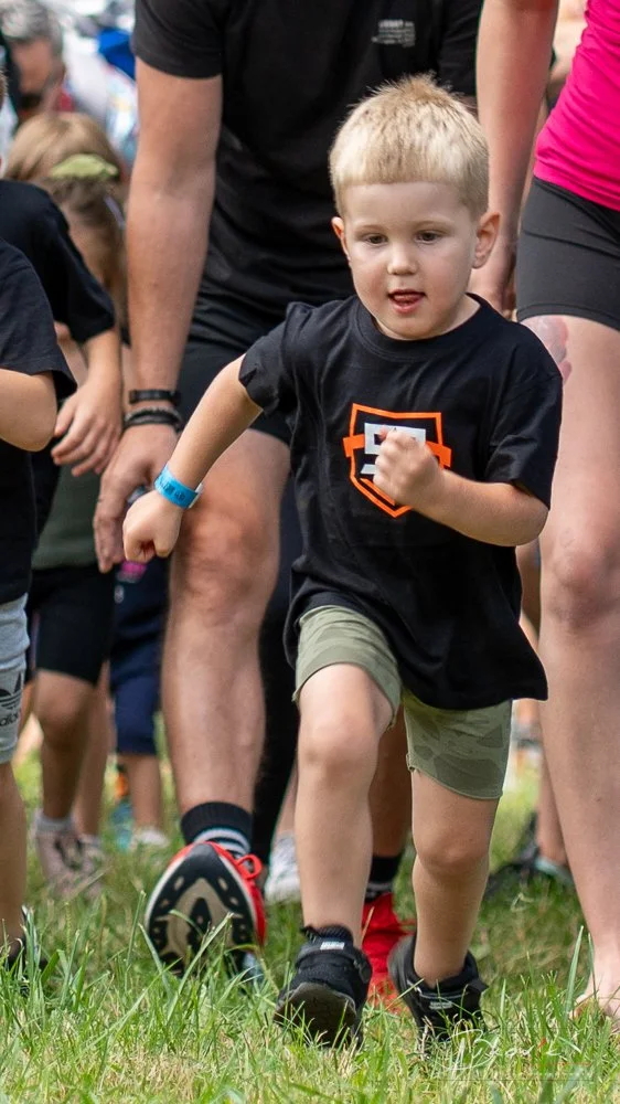 Young boy running in a race with other children and adults around him on a grassy field.